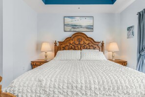 Ein ordentlich bezogenes Bett mit weißer Steppdecke, zwei Nachttische mit Lampen, ein Holz-Kopfteil mit aufwendigen Schnitzereien und ein Vogelbild an der Wand.