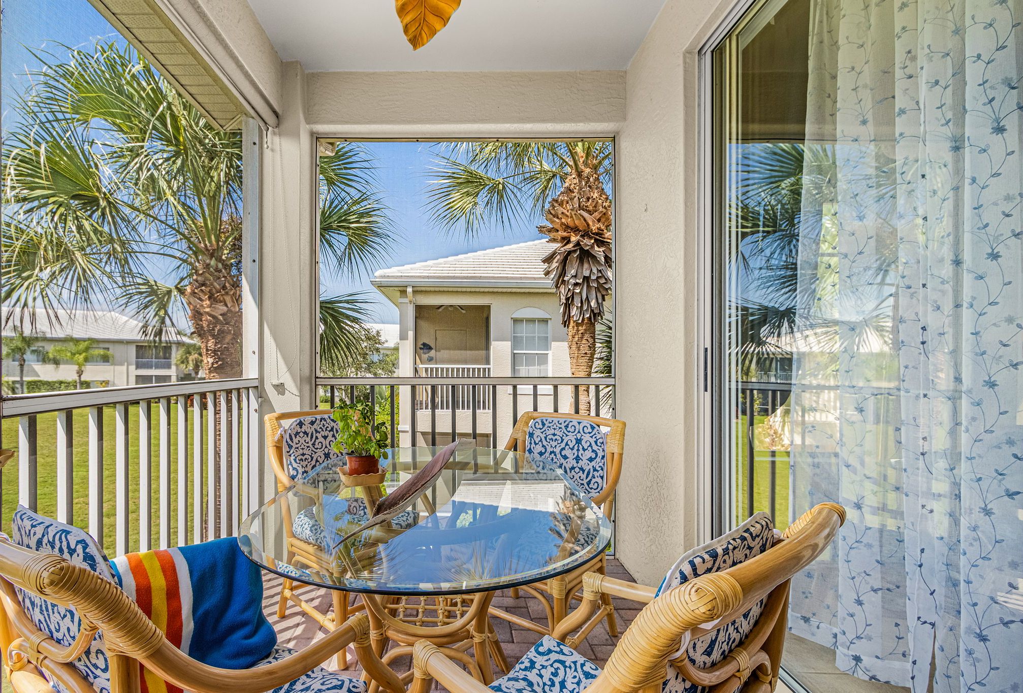 Un balcón con mobiliario de mimbre, una mesa de vidrio y vista a una casa y palmeras.
