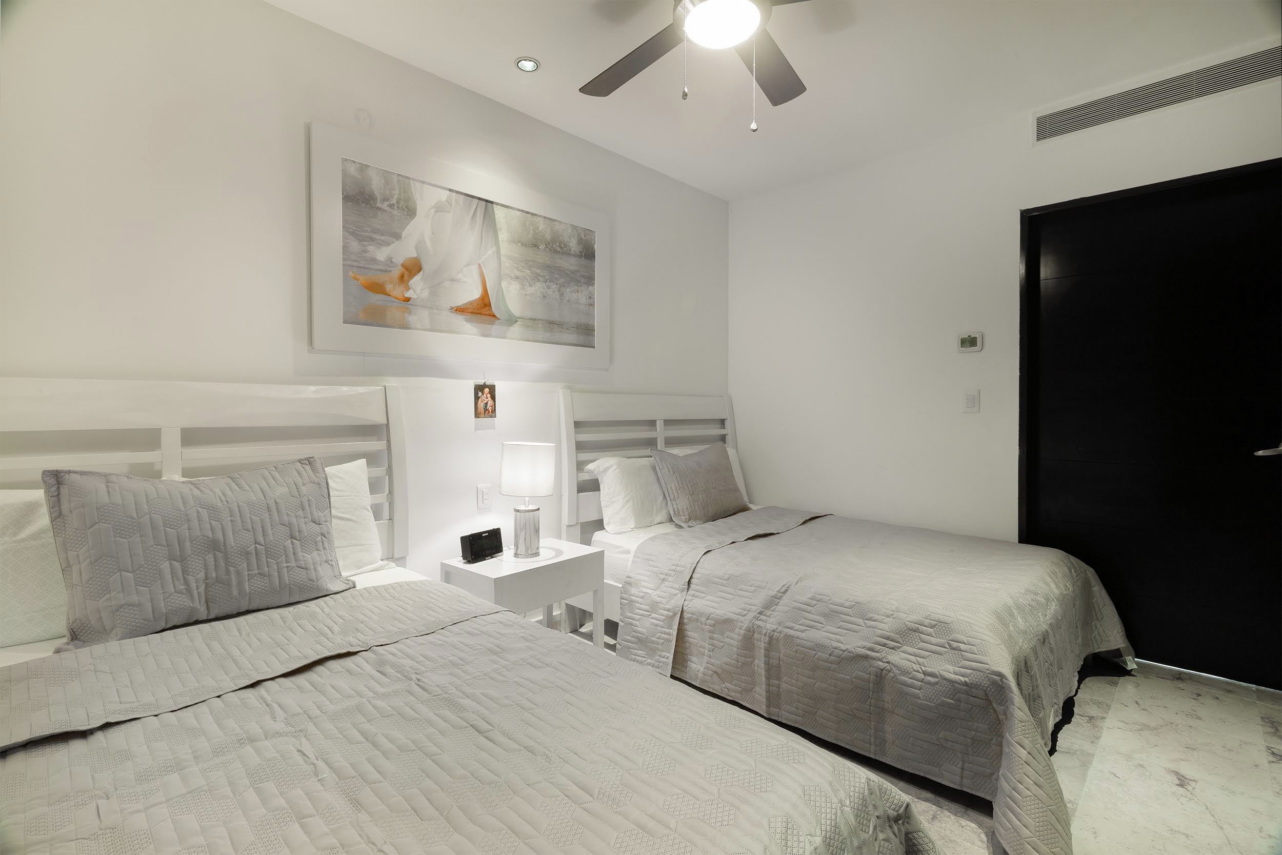Un dormitorio con dos camas, una mesita de noche, un ventilador de techo y un cuadro en la pared.
