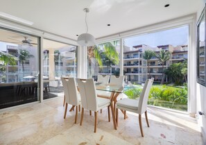 A modern dining area with a glass table and white chairs, large windows offering a view of a pool and surrounding buildings. - Ocean View / Fitness Center / Private Beach / Huge Pool (Playa Del Carmen)