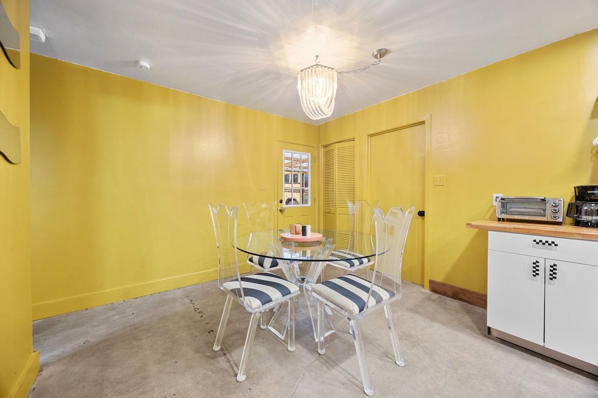 A dining area with a glass table and chairs, a chandelier, a kitchenette with a toaster, and a yellow wall.
