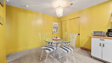 A dining area with a glass table and chairs, a chandelier, a kitchenette with a toaster, and a yellow wall.