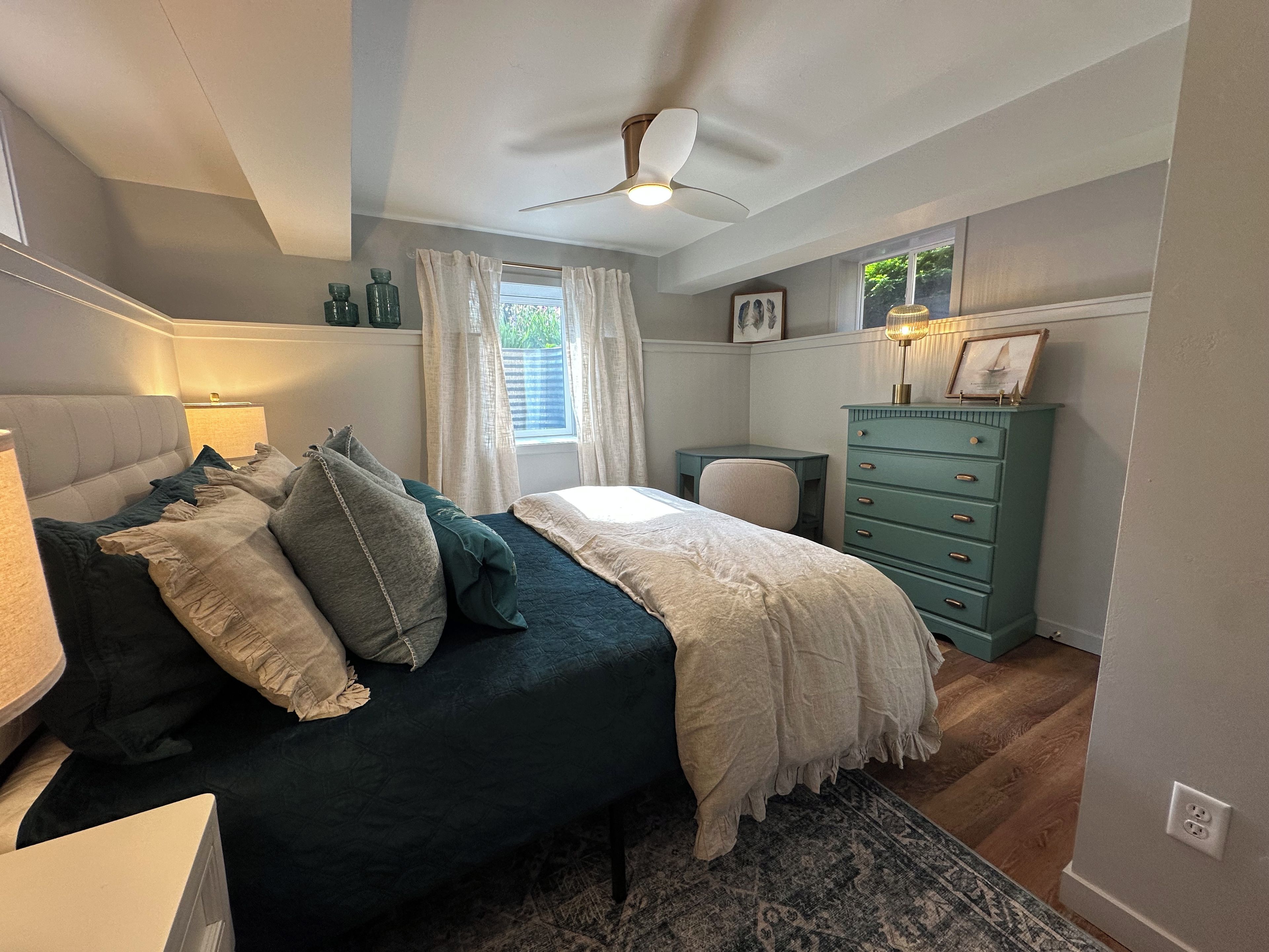 A bedroom with a bed, a desk, a dresser, a window with curtains, and a ceiling fan.