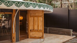 Una estructura de madera con un toldo verde estampado, una puerta de madera y un espacio interior visible con una mesa y sillas.