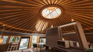 Interior de una yurta con tragaluz circular, vigas de madera, zona de comedor y televisor.