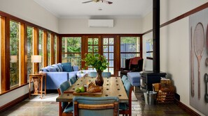 A room with a large wooden dining table, blue upholstered furniture, and a view of greenery through large windows. - Warburton Stays | Grand View (Warburton)