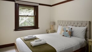 A bedroom with a bed, a nightstand with a lamp, a window with a view of trees, and a bedside table with towels. - Warburton Stays | Grand View (Warburton)