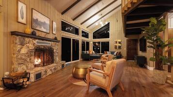 A spacious living room with a stone fireplace, wooden beams, and large windows.