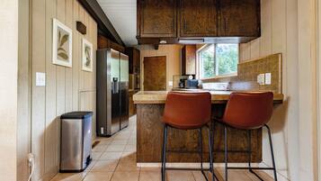 A kitchen with wooden cabinets, a stainless steel refrigerator, a trash bin, and two bar stools.