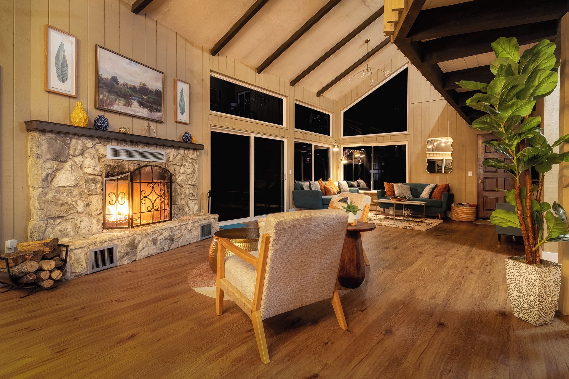 A cozy living room with a stone fireplace, wooden flooring, and large windows.