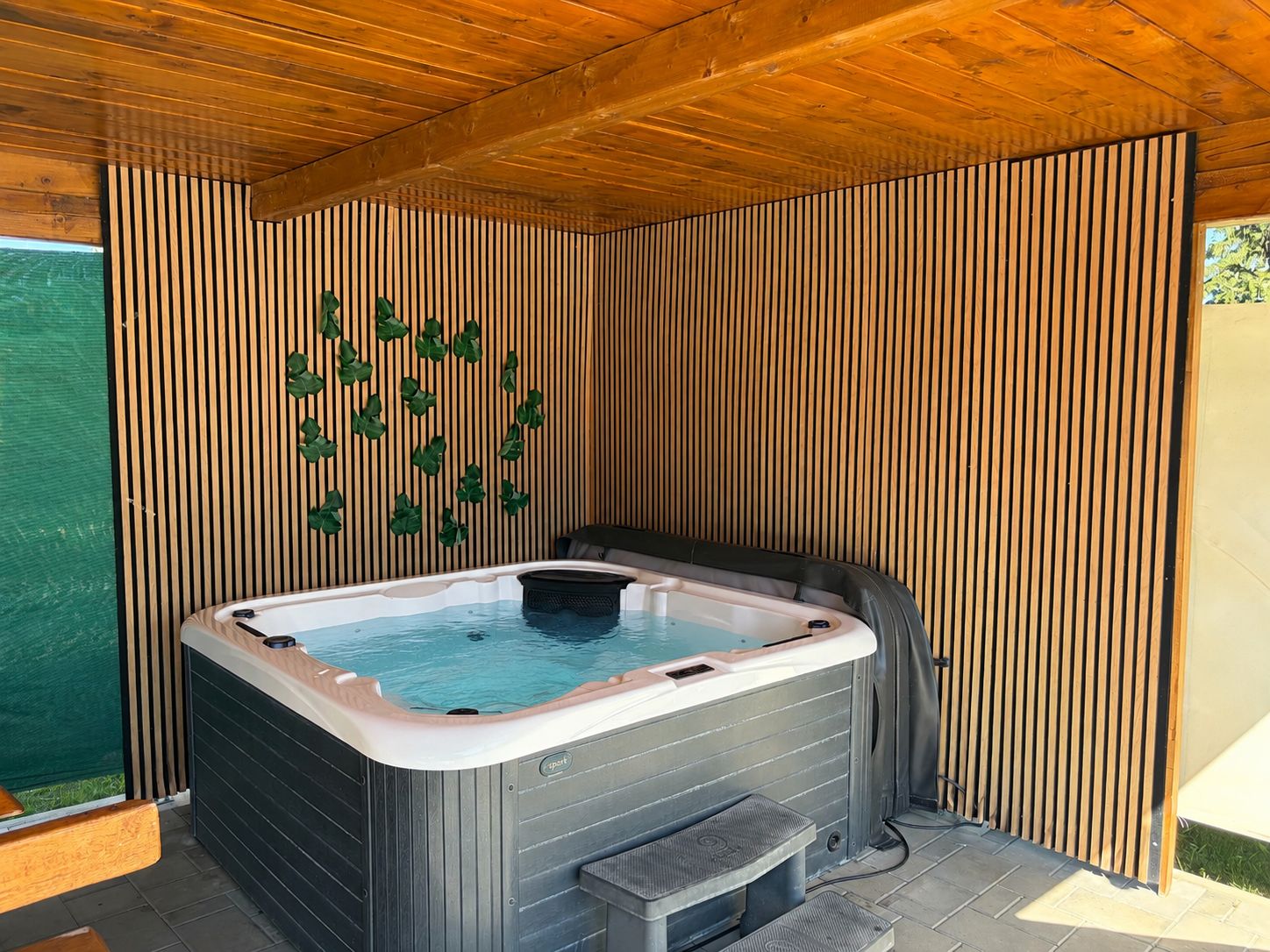 A hot tub in a covered outdoor area with vertical wooden slats on the walls and a bench to the left.