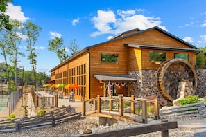 A wooden building with a large water wheel, outdoor seating, and a stone wall. - 3-Level Luxury Cabin 🌲 Creek Views, Hot Tub & Top-Tier Resort Pools! (Hollister)