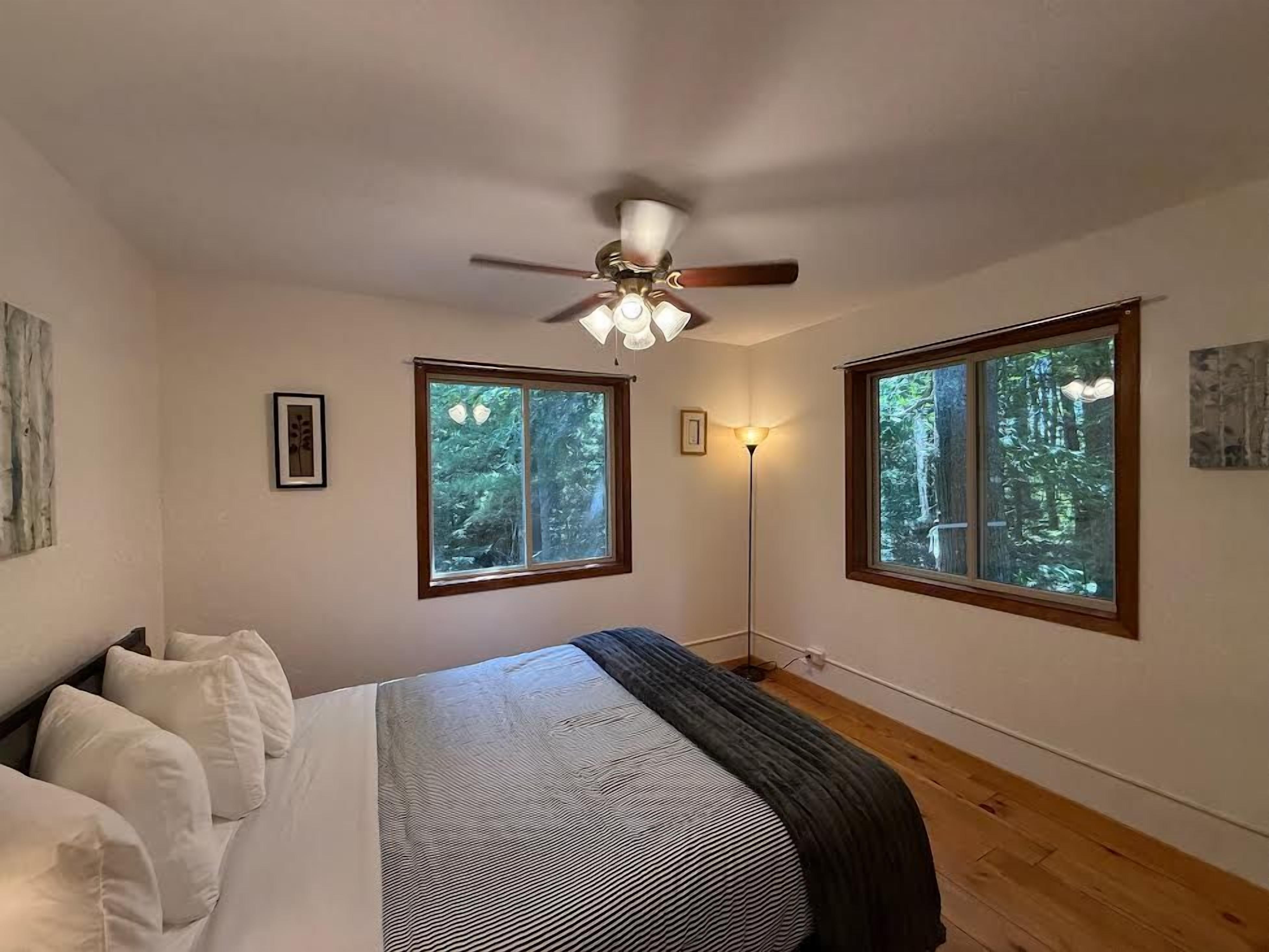 Une chambre à coucher avec un lit, un ventilateur de plafond, deux fenêtres donnant sur des arbres et une lampe sur pied.