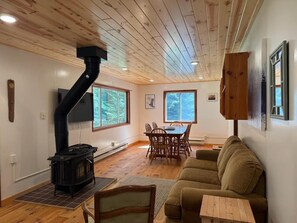 A cozy living room with a wood-burning stove, a dining area with chairs, and a sofa. - Turtle Cabin – Gichigami Island Properties 2 Bedrooms • 1 Bathroom • Sleeps 4 • Beach Access (La Pointe)