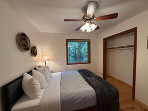 A bedroom with a bed, a ceiling fan, a window with a view of trees, and a walk-in closet. - Turtle Cabin – Gichigami Island Properties 2 Bedrooms • 1 Bathroom • Sleeps 4 • Beach Access (La Pointe)