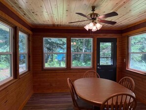 Ein Innenraum einer Holzhütte mit einem Esstisch, Stühlen, einem Deckenventilator und großen Fenstern.