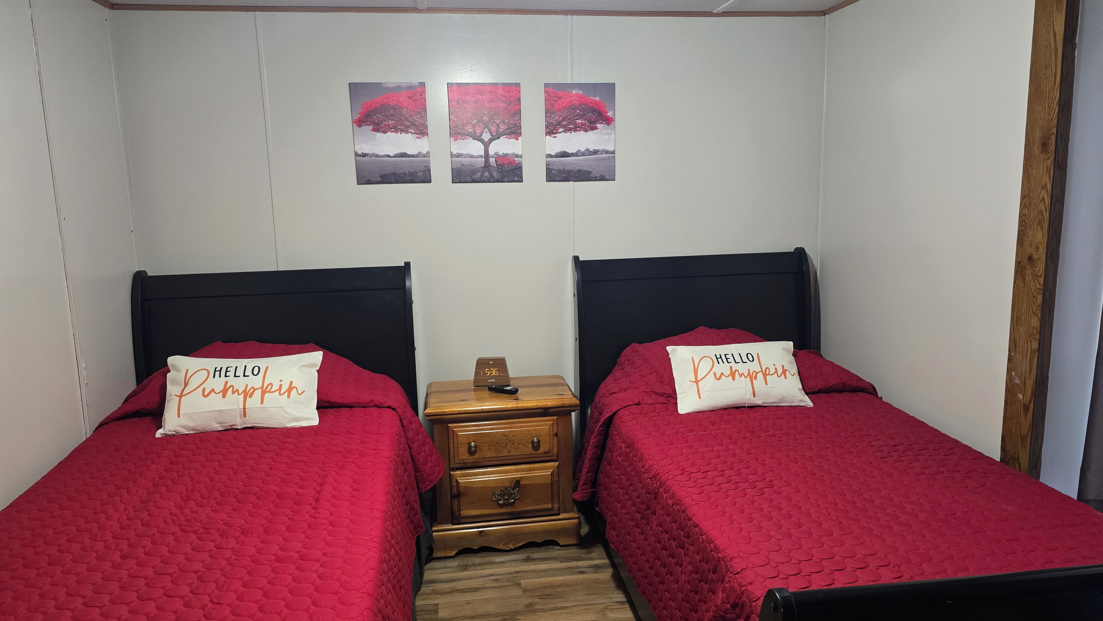 A bedroom with two beds, each with a red bedspread and a pillow with 'Hello Pumpkin' text. There is a wooden nightstand between the beds with a clock and a remote control.