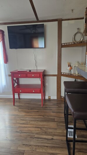 A room with a wooden floor, a red desk with drawers, a television mounted on the wall, and a brown chair.