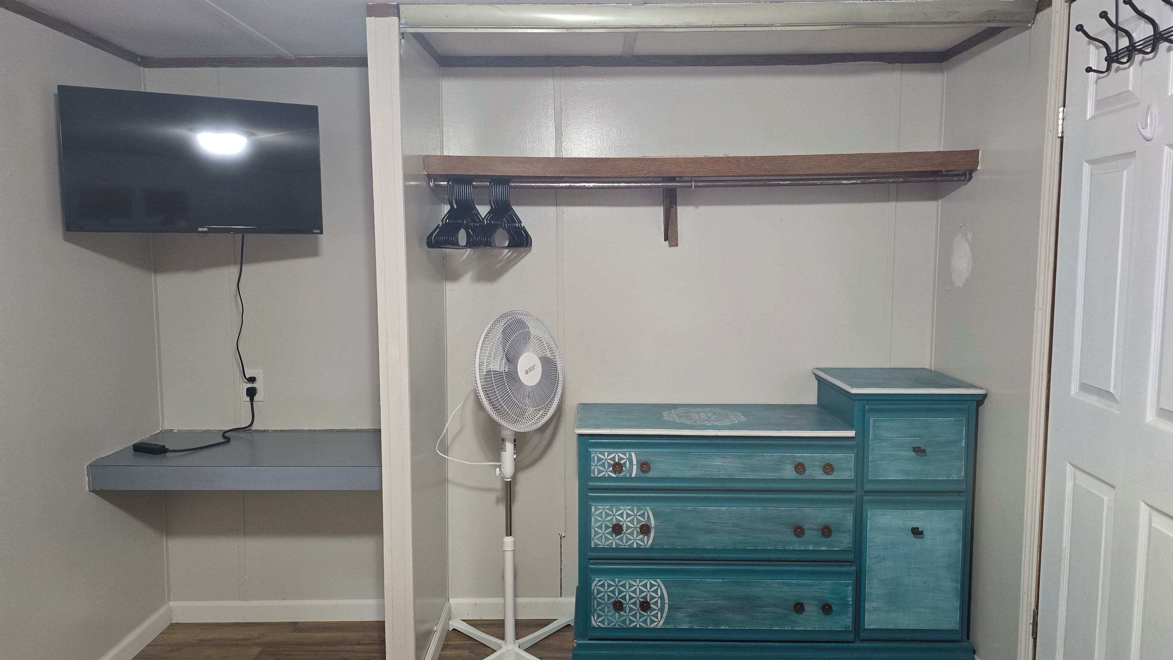 A room with a flat-screen TV mounted on the wall, a standing fan, a teal dresser with drawers, and a shelf above the dresser.
