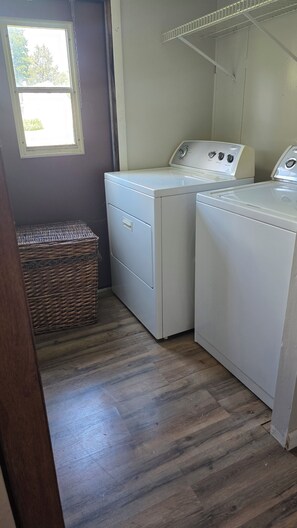 A laundry room with two white washers and dryers, a wicker basket, and a window with a view of trees. - Hello Pumpkin!  2-bedroom mobile home in Caseyville, IL (Caseyville)