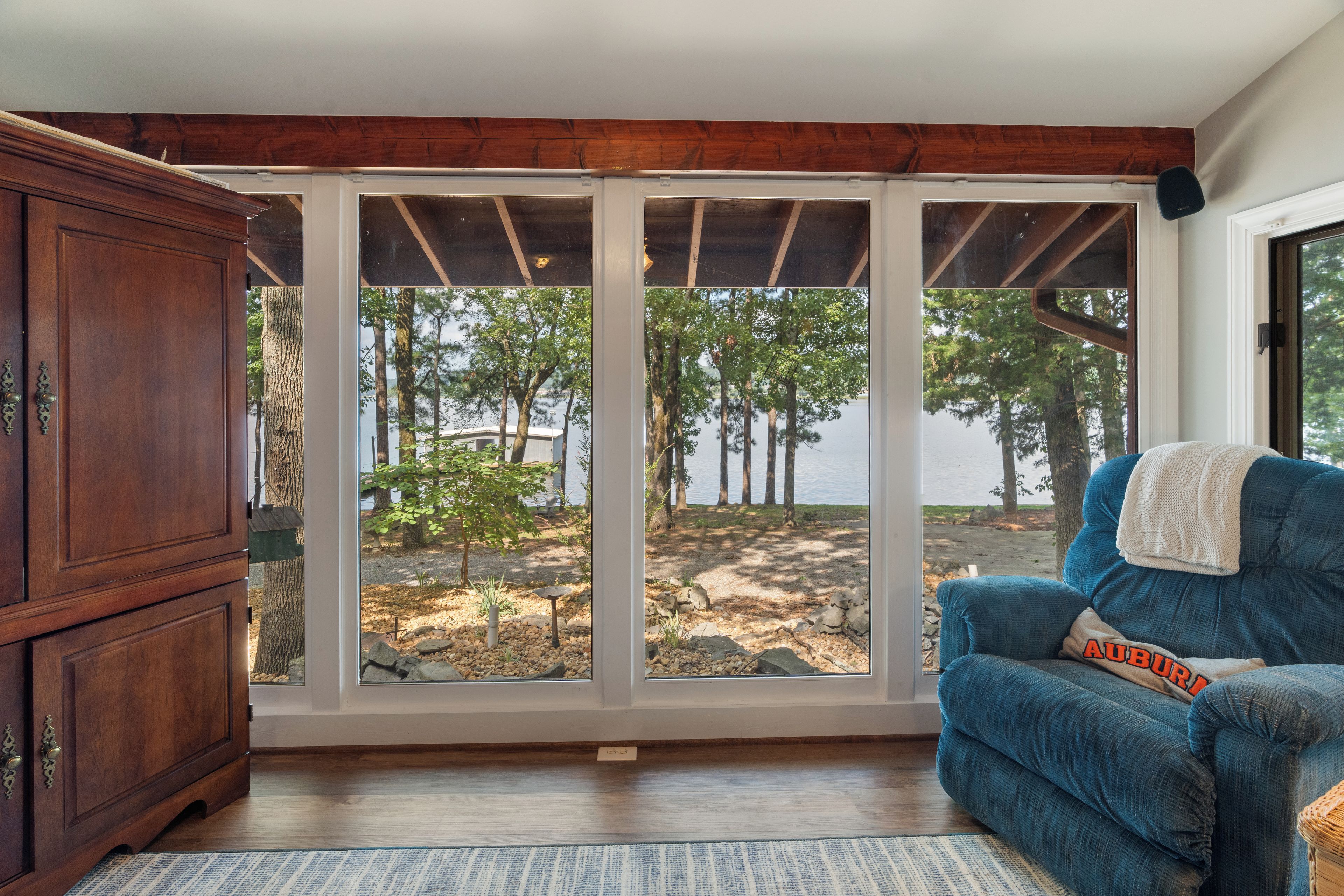A room with a large sliding glass door leading to a wooded area with a view of a body of water. There is a blue sofa with a cushion labeled 'AUBURN' and a wooden cabinet to the left.