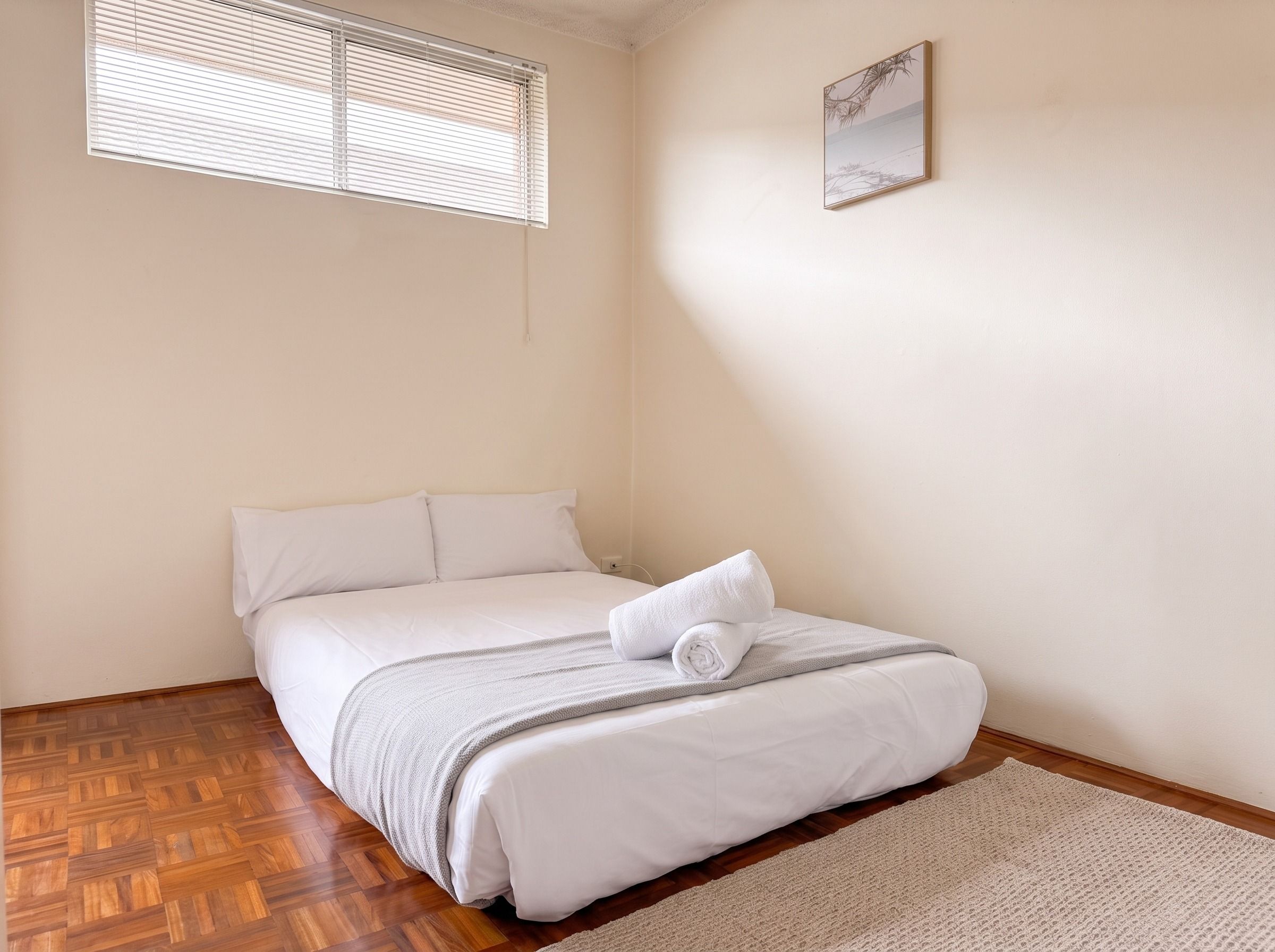Une chambre à coucher avec un lit, une fenêtre avec des stores, un tableau accroché au mur et un plancher en bois.