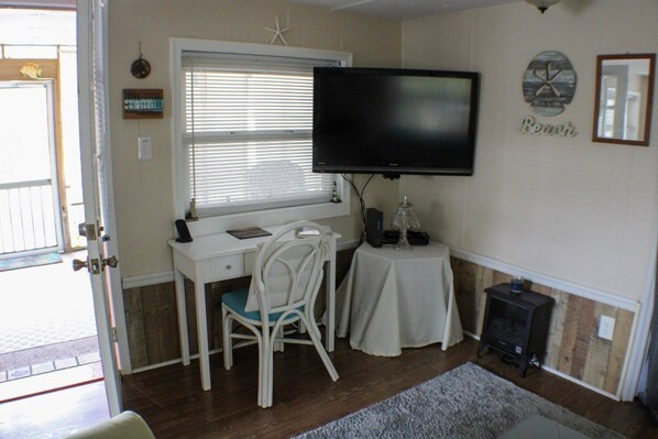 A room with a desk, chair, television, and a small table with a lamp.