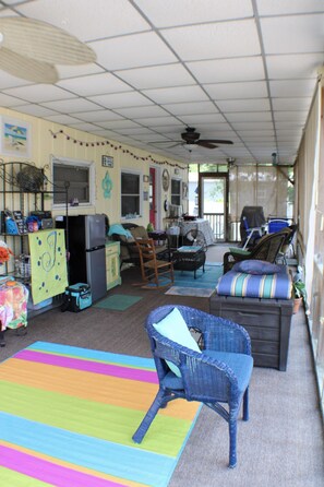 A covered outdoor living space with a ceiling fan, a refrigerator, a dining table, and various seating arrangements including a rocking chair and a sofa.