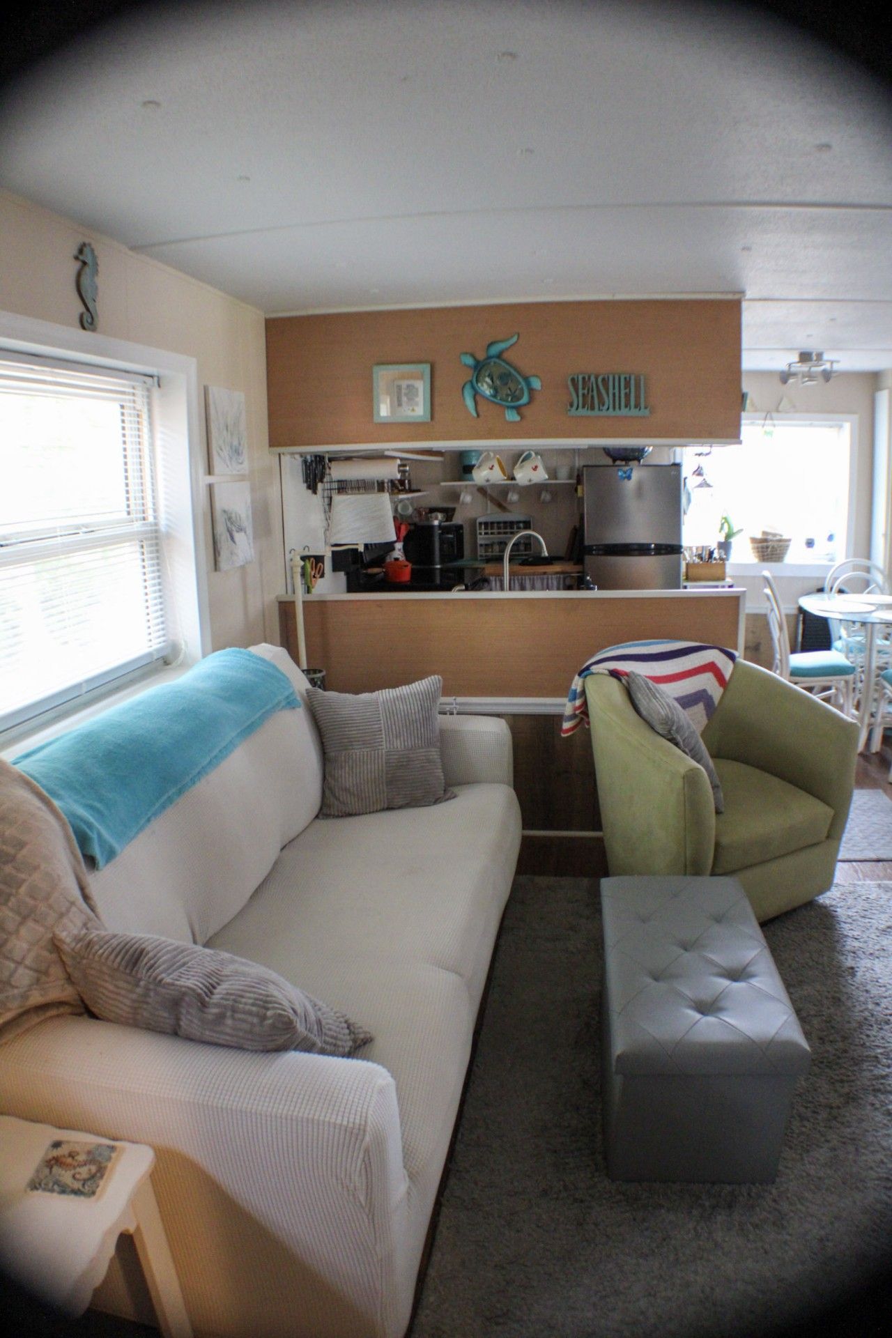 Un espace de vie compact comprenant un canapé, un fauteuil vert, un petit pouf, une kitchenette et une fenêtre avec des stores.