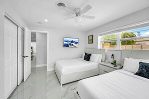 Ein modernes Schlafzimmer mit zwei Betten, einem Deckenventilator, einem Fernseher und einem Fenster mit Blick auf Grünflächen.
