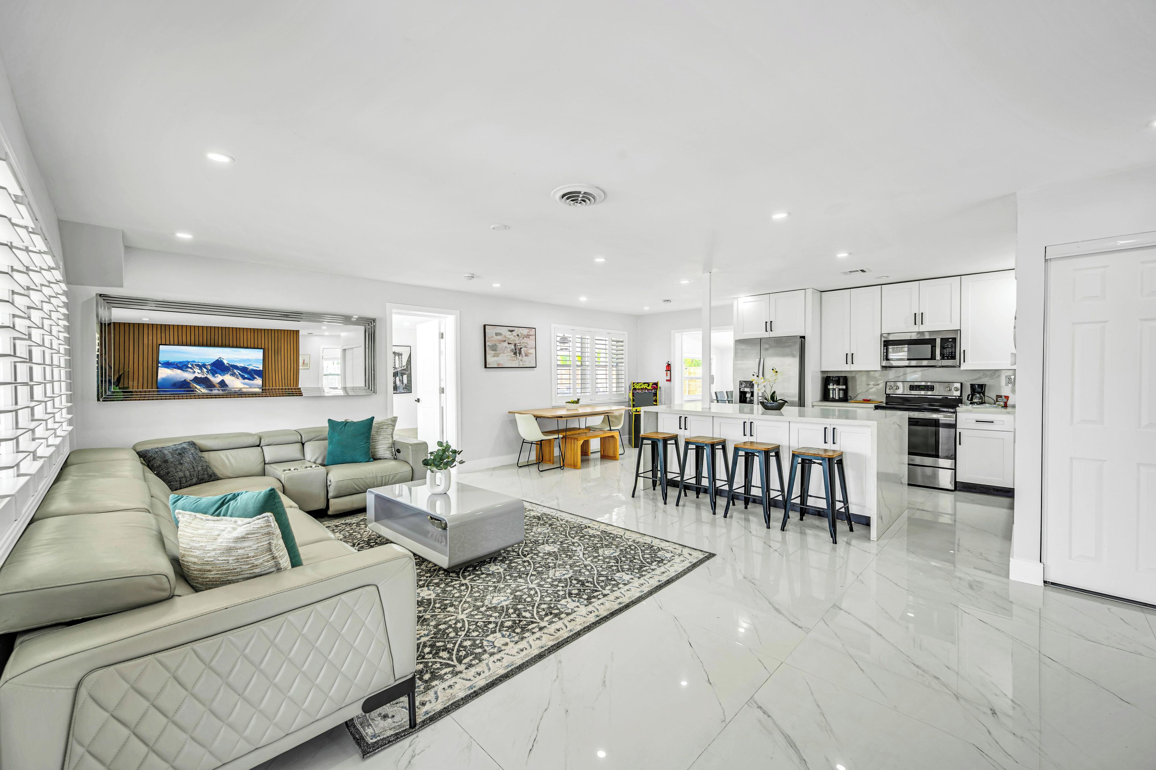 A modern living room with a sofa, coffee table, and a TV. It leads to a kitchen with a dining area and bar stools.