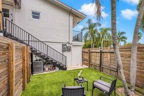 Ein Wohngrundstück mit Holzzaun, einer Treppe zu einem Balkon, einer kleinen Terrasse mit Tisch und Stühlen sowie einem Rasen.