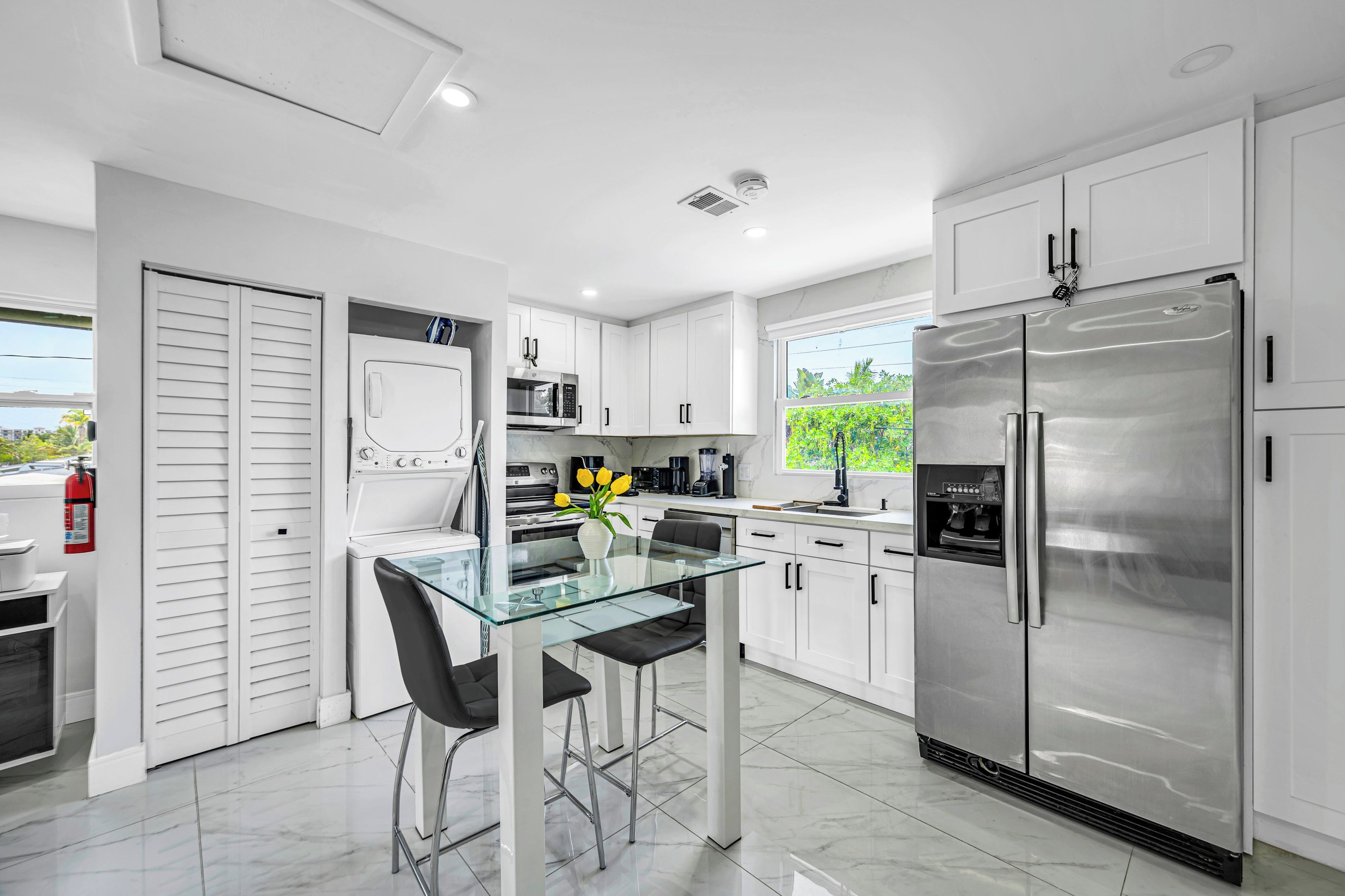 A modern kitchen with white cabinets, a stainless steel refrigerator, a glass dining table, and a coffee machine.