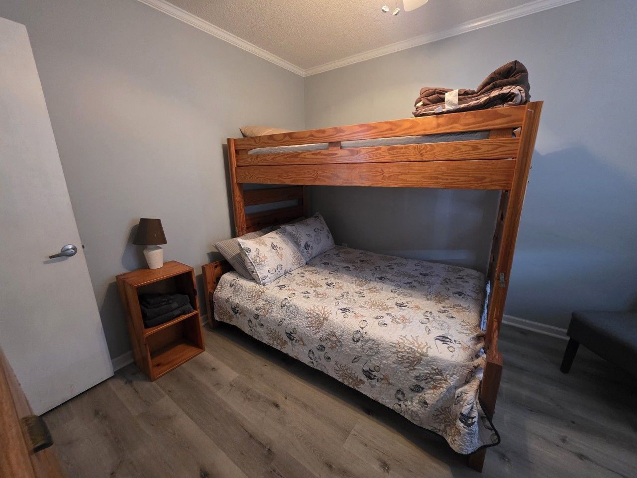 A bunk bed with a patterned bedspread, a bedside table with a lamp, and a small shelf unit.