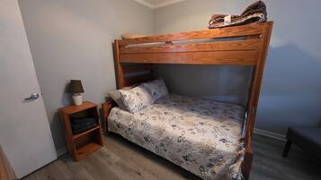A bunk bed with a patterned bedspread, a bedside table with a lamp, and a small shelf unit.