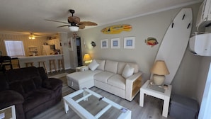 A living room with a sofa, a coffee table, a ceiling fan, a surfboard decoration, and a kitchen area in the background. - Beach Retreat Oceanview Balcony (North Topsail Beach)