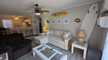 A living room with a sofa, a coffee table, a ceiling fan, a surfboard decoration, and a kitchen in the background.