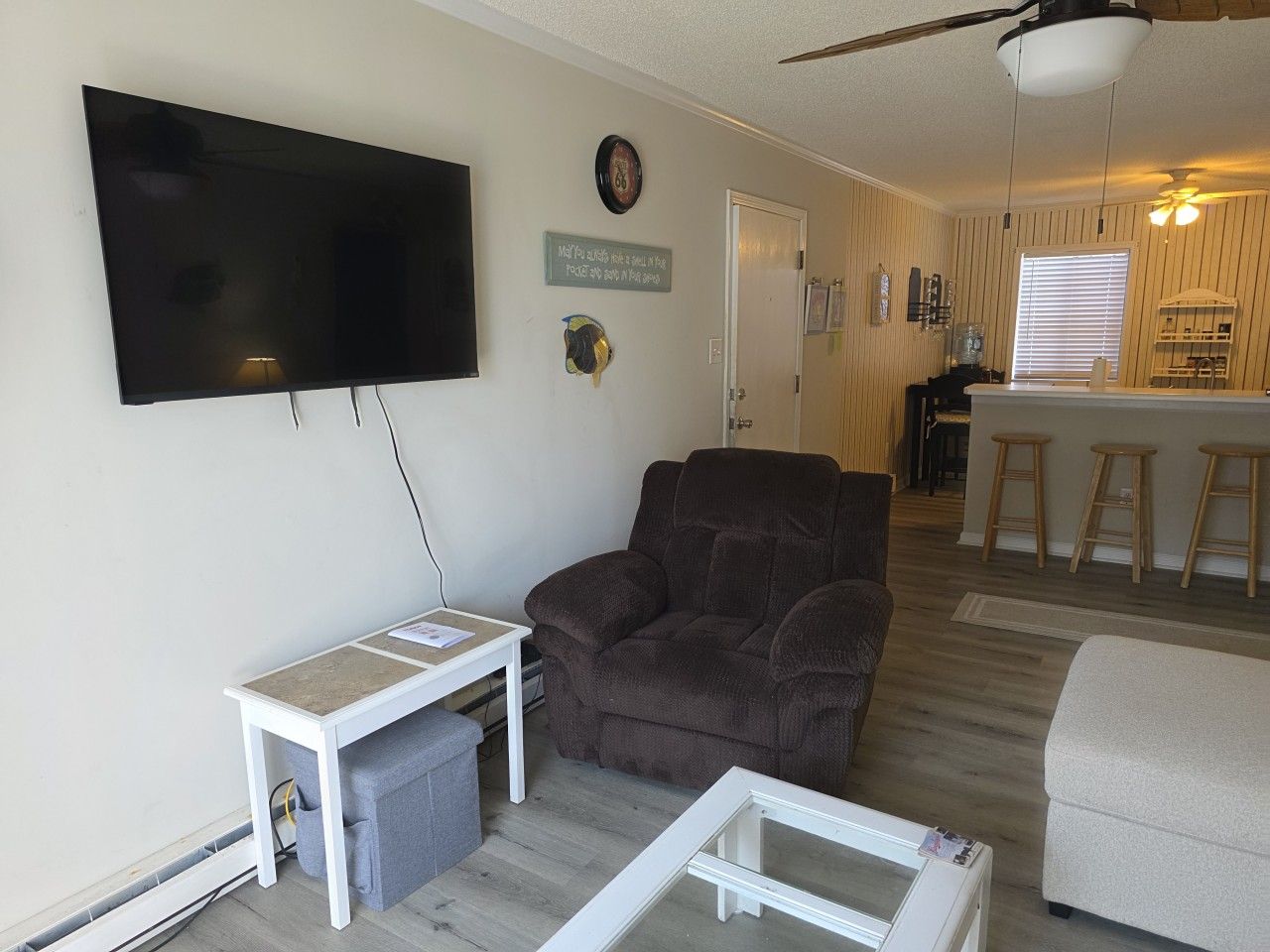 A living room with a flat-screen TV mounted on the wall, a brown sofa, a small white table, and a glass-top coffee table.