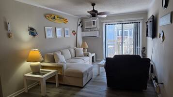 A living room with a sofa, ottoman, side tables, a ceiling fan, and a window with blinds.
