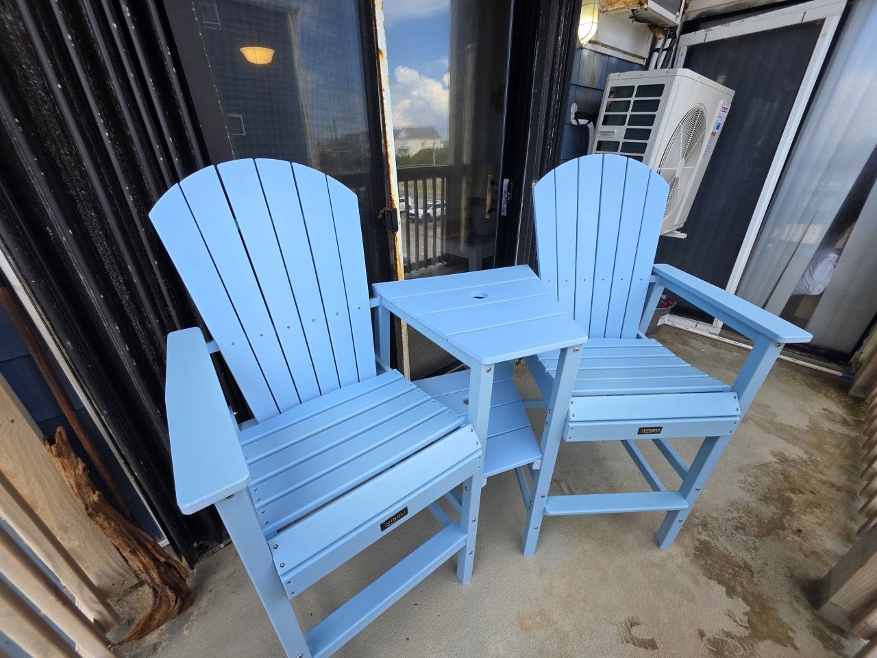 Zwei blaue Adirondack-Stühle und ein kleiner Tisch vor einem Gebäude mit einer schwarzen Tür und einer Klimaanlage.