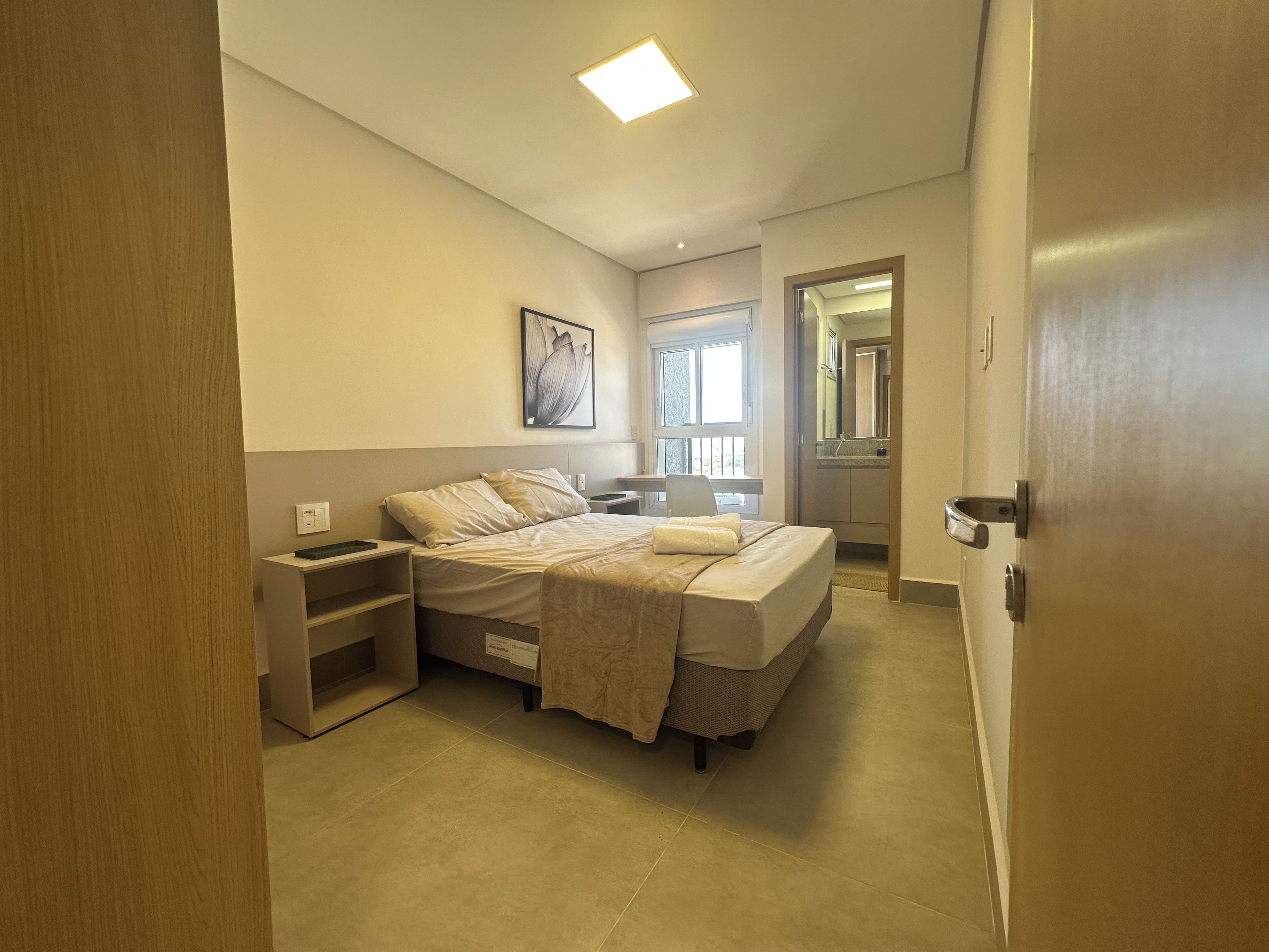 A hotel room with a bed, bedside tables, a desk, a window, and a bathroom visible through an open door.