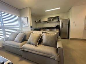 A modern living room with a grey sofa, a kitchen area with appliances, and a window with blinds.