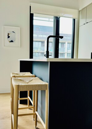 Una cocina moderna con encimera negra, ventana y dos taburetes de fibras naturales.