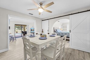 A dining area with a white table set for four, a ceiling fan, and a view into a room with a bed and a TV. - Poolside Escape for Family Fun in Sunny Cape Coral Getaways - Country Club Villa - Roelens (Cape Coral)