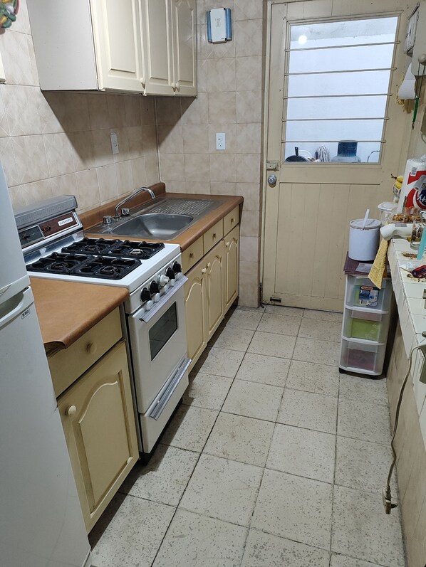 A compact kitchen with a gas stove, oven, and sink. There is a window with horizontal blinds, and a small storage unit with containers. - Extraordinary house in Los Pinos, Morelia (wheelchair accessible). (Morelia)