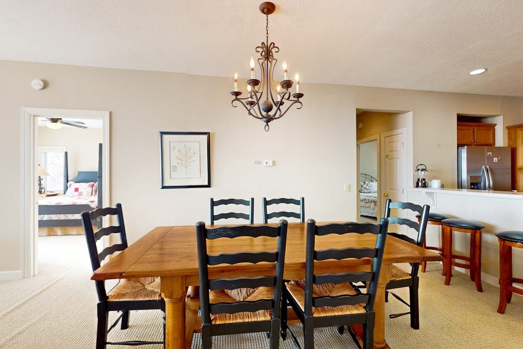 A dining room with a wooden table and chairs, a chandelier, and a kitchen in the background.