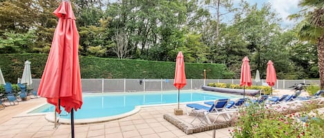 Zona de piscina con sillones de descanso y sombrillas, rodeada por una valla y follaje.