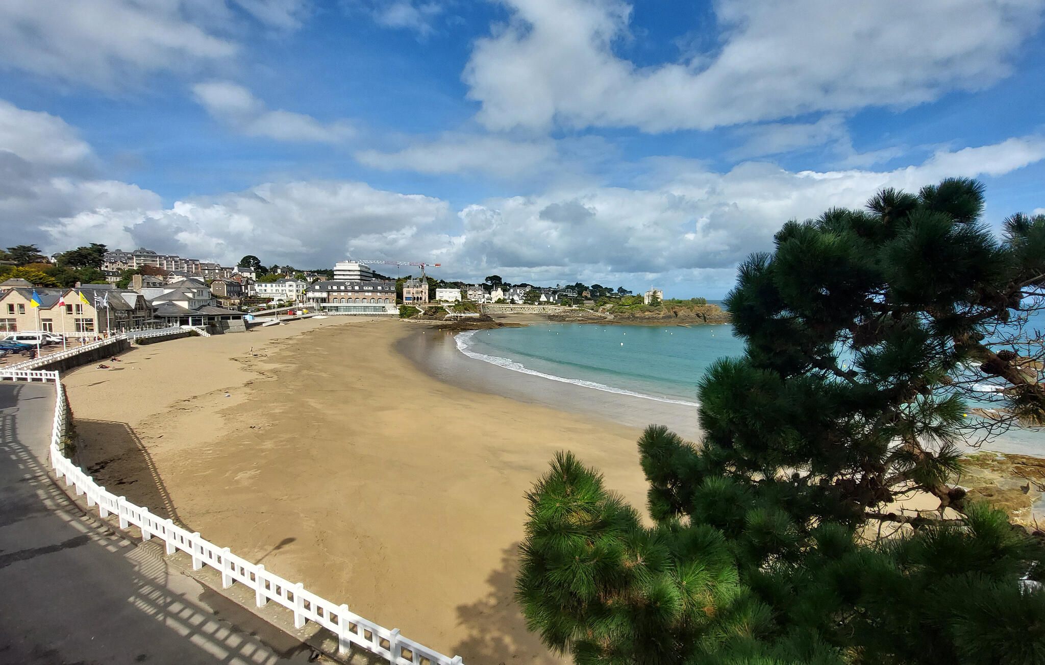 A coastal beach with a white fence, buildings in the background, and a clear sky.
