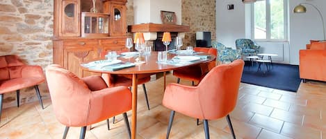 Un comedor con una mesa redonda dispuesta para cuatro personas, rodeada de sillas color naranja, en una habitación con paredes de piedra y un amplio ventanal.