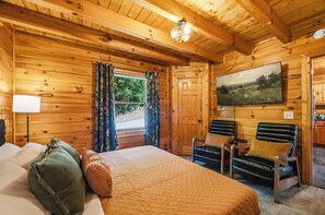 A wooden cabin room with a bed, two chairs, a painting, and a window with curtains. - Firefly Forest • Stunning Mtn Views • Hot Tub • Sauna • Firepit • 5 Min to Pkwy! (Pigeon Forge)